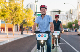 Two people wearing helmets ride bikes in a city bike lane on a sunny day, with trees and buildings in the background. The person in front is smiling at the camera.