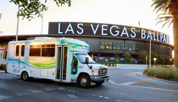 A small shuttle bus with green and blue accents drives past Las Vegas Ballpark, which has large white letters on its exterior. Palm trees and landscaping are visible in the foreground.