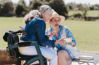 Two women sitting outside together, one in a wheelchair and the other on a bench, looking at something in a notebook or card and smiling, with a grassy field in the background.