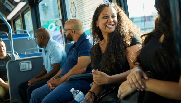 A woman smiles while sitting on a bus, holding a water bottle. Other passengers are seated around her, engaged in conversation or looking out the window. Sunlight streams through the windows, creating a warm atmosphere.