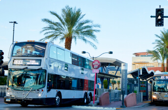 A silver double-decker public bus stops at a modern outdoor bus station lined with palm trees, with another bus in the background and a tall building visible behind them.