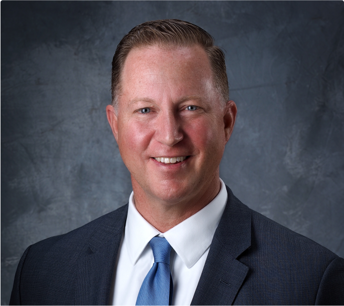 Frame 124 A man in a suit and blue tie smiles at the camera against a gray textured background. He has short, neatly styled hair and a light complexion.