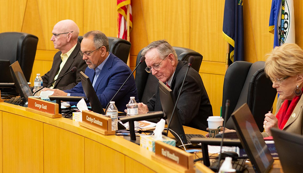RTC-Board-May2019-3483_web 1 Four people sit at a long desk in a formal meeting room with laptops, nameplates, documents, and flags behind them. They appear focused, reading or writing notes during a session.