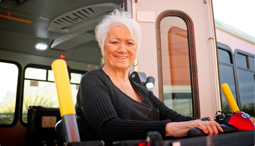 S.A.F.E. Program An older woman with white hair smiles while sitting in a wheelchair lift outside a bus, preparing to board. Bright yellow safety bars and the bus entrance are visible behind her.