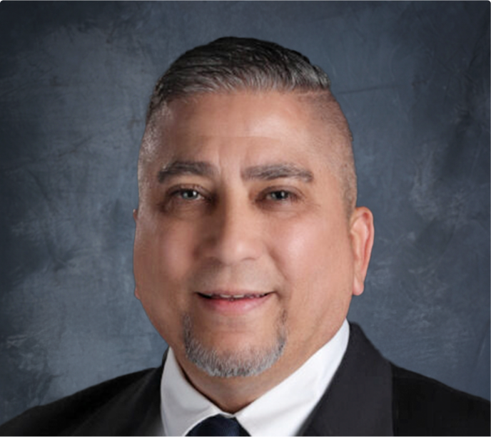 image-1 A middle-aged man with short, neatly styled dark hair and a goatee, wearing a suit and tie, smiles at the camera against a dark blue textured background.
