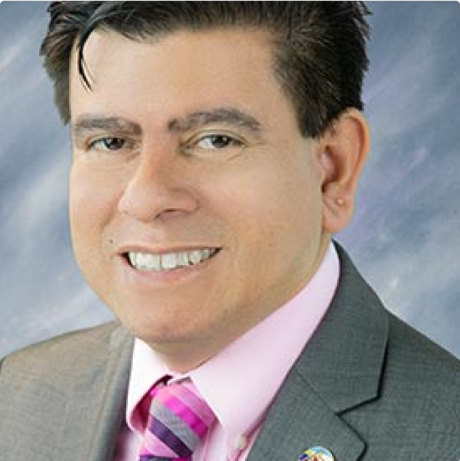 image-1 A man with short dark hair wearing a gray suit, pink shirt, and striped pink tie smiles at the camera against a soft, blurred background.