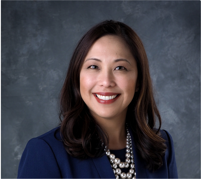 image Professional portrait of a woman with long dark hair wearing a navy blazer, white blouse, and layered pearl necklace, smiling in front of a gray textured background.