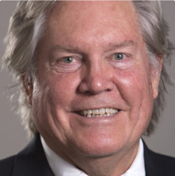 image-2 A smiling older man with gray hair wearing a dark suit jacket and white shirt, shown in a close-up portrait against a neutral background.