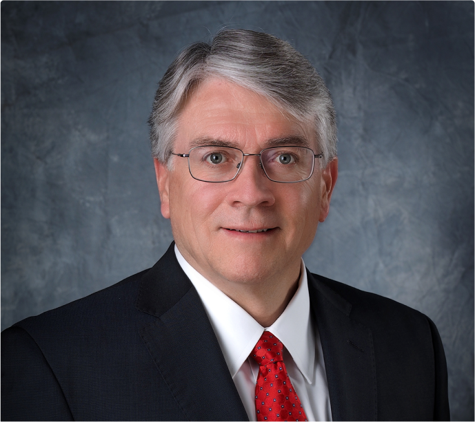 image-2 A middle-aged man with gray hair and glasses wearing a dark suit, white shirt, and red tie, posing in front of a mottled blue-gray background.