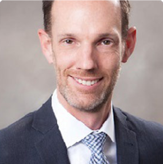 image A man with short brown hair and a trimmed beard, wearing a dark suit, white shirt, and patterned tie, smiles at the camera against a plain, light gray background.
