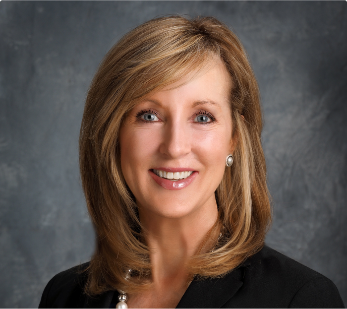 image A woman with shoulder-length blonde hair, wearing a black blazer and pearl necklace, smiles in front of a gray background.