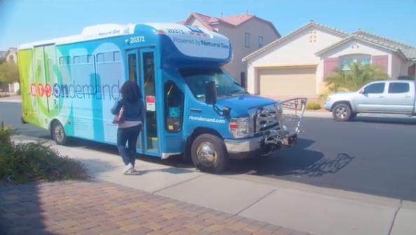 276979f1f06efe42f920934b1fd1cb8a02923fa6 A woman walks toward a blue and green shuttle bus labeled on demand parked at the curb in a suburban neighborhood with houses and a clear sky in the background.