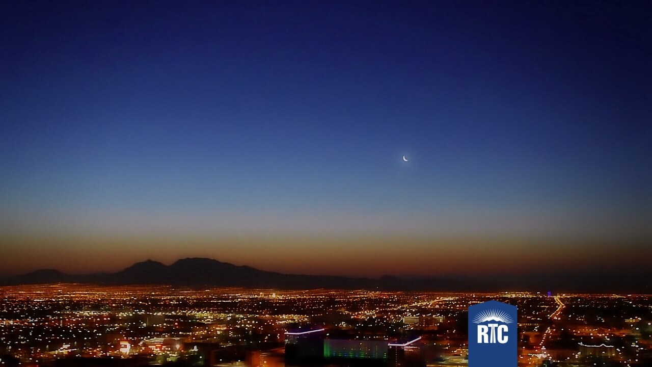 A cityscape at dusk with a crescent moon in a darkening sky, city lights glowing below, and distant mountains on the horizon. An RTC logo is in the bottom right corner.