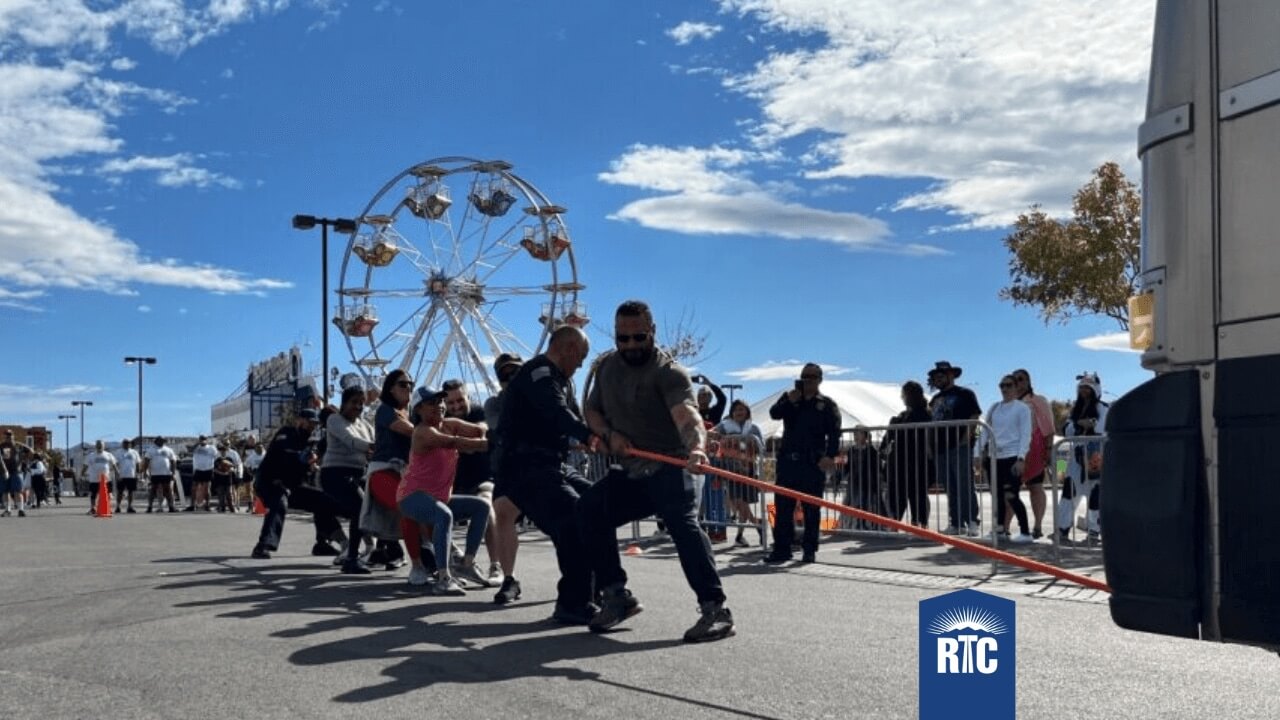 People participate in a team tug-of-war, pulling a rope attached to a large vehicle, with a Ferris wheel and crowd in the background under a blue sky. RTC logo appears in the bottom right corner.